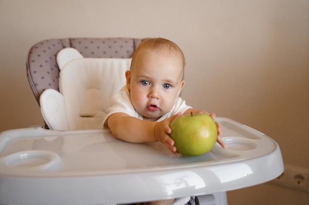 een kind op een kinderstoel in een wit rompertje speelt met een groene appel Eten