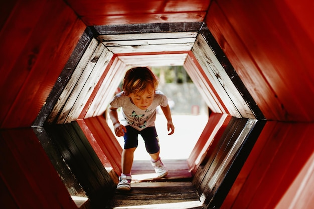 Een kind speelt in een avontuurlijk park door een rode houten tunnel.