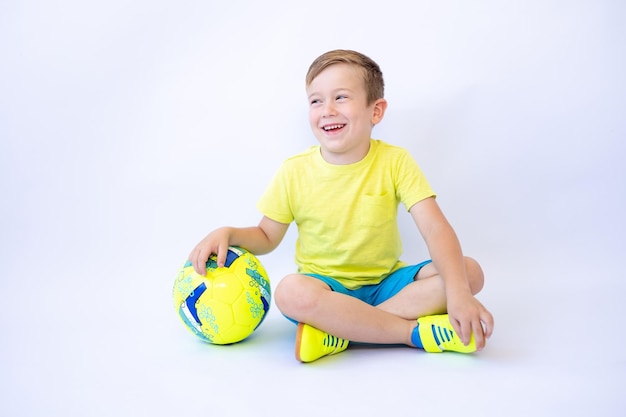 Een kindjongen in sportkleding zit op een witte achtergrond met een voetbal in zijn handen en sport een gezonde levensstijl