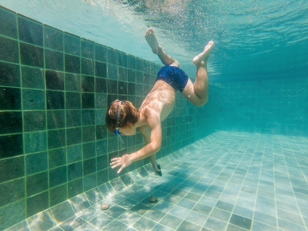 Een kindjongen zwemt onder water in een zwembad, glimlachend en adem inhoudend, met een zwembril.