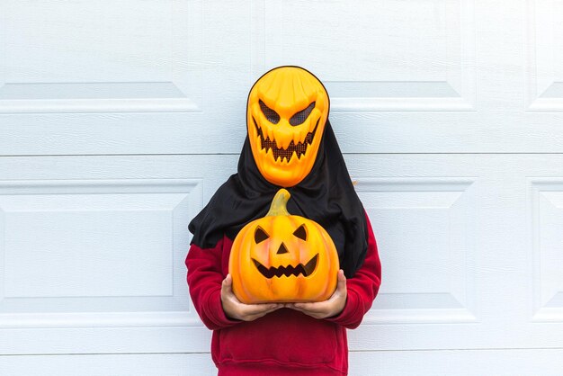 Een kindmeisje dat een eng Halloween-pompoenmasker draagt en een pompoen over een witte garagedeur houdt Concept van feestkostuum carnaval terreurangst en herfst