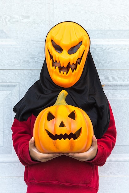 Foto een kindmeisje dat een eng halloween-pompoenmasker draagt en een pompoen vasthoudt die met haar hand over een witte garagedeur schrikt concept van feestkostuum carnaval terreur angst en herfst