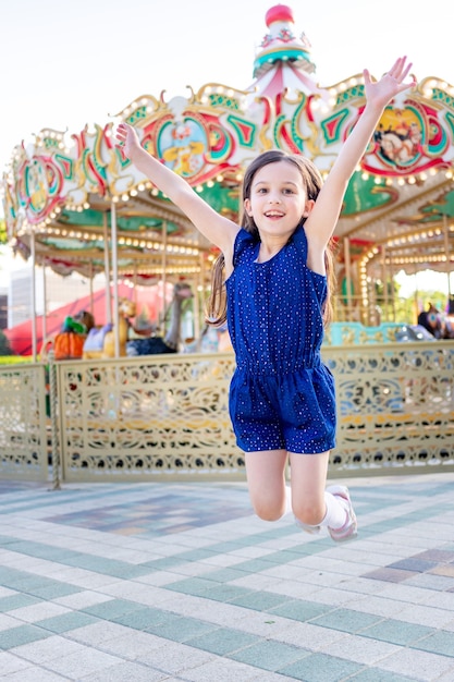 Een kindmeisje in een pretpark in de zomer springt van geluk rond de carrousels en lacht, het concept van zomervakantie en schoolvakanties