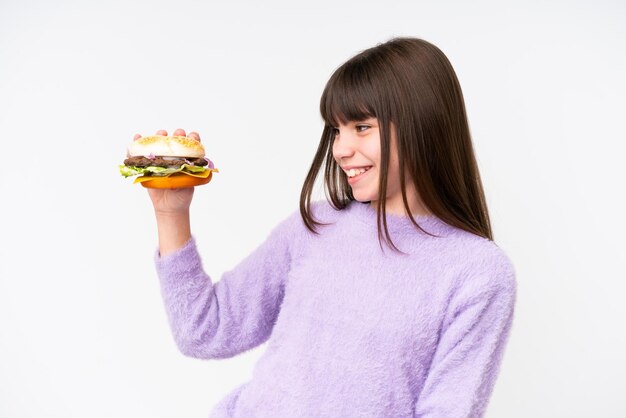 Een klein blank meisje dat een hamburger vasthoudt over een geïsoleerde achtergrond met een gelukkige uitdrukking