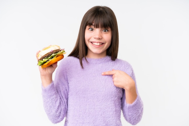 Een klein blank meisje dat een hamburger vasthoudt over een geïsoleerde achtergrond met een verrassende gezichtsuitdrukking