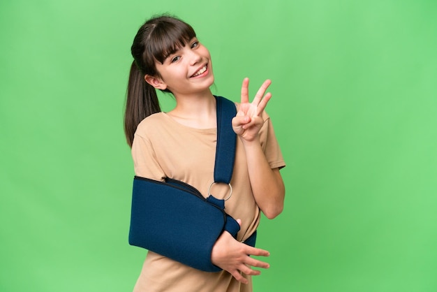 Een klein blank meisje met een gebroken arm en een draagdoek over een geïsoleerde achtergrond, gelukkig en drie tellend met vingers