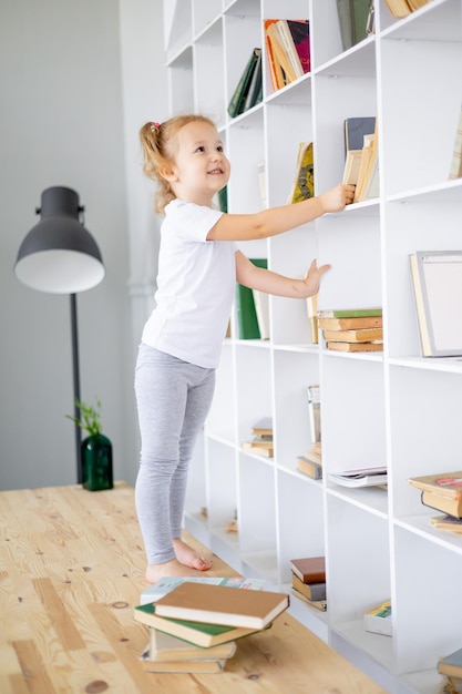 Een klein blond meisje haalt een boek tevoorschijn in een heldere bibliotheek waar ze thuis lesgeeft en boeken voor kinderen leest