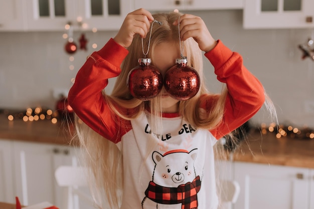 Een klein blond meisje in een rode kerstpyjama zit in de keuken en houdt twee rode kerstballen in haar handen waarachter ze haar gezicht verbergt. ruimte voor tekst. hoge kwaliteit foto