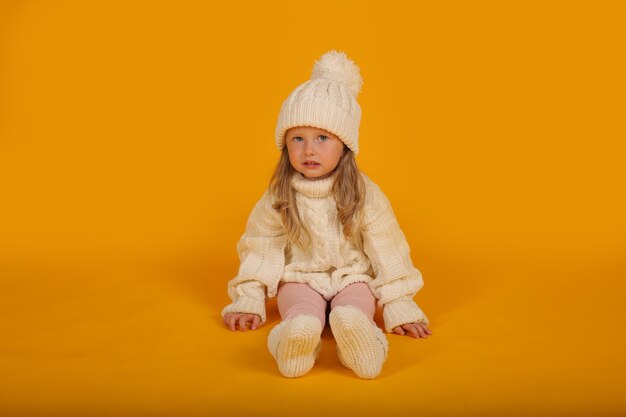 een klein blond meisje in een witte gebreide trui een witte muts en gebreide sokken