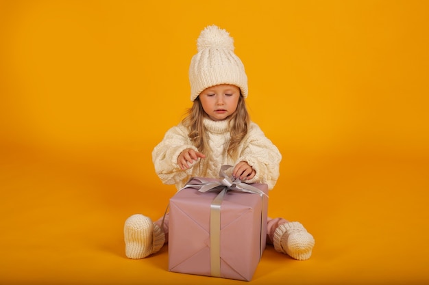 een klein blond meisje in een witte gebreide trui, muts en sokken met een geschenkdoos in haar handen