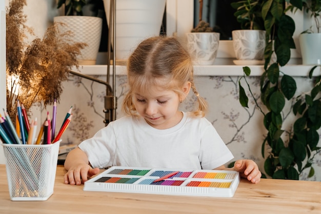 Een klein blond meisje met een helder, kleurrijk palet van gekleurde plasticine om aan tafel te modelleren