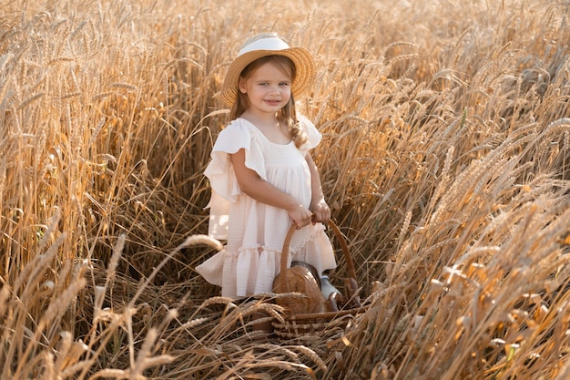 Een klein blond meisje met een strohoed houdt een mand met brood in een tarweveld, milieuvriendelijke landbouwproducten