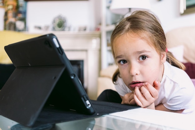 Een klein blond meisje voor een tablet die naar de camera in haar woonkamer kijkt concept van kindertechnologie die plezier heeft met internet en connectiviteit