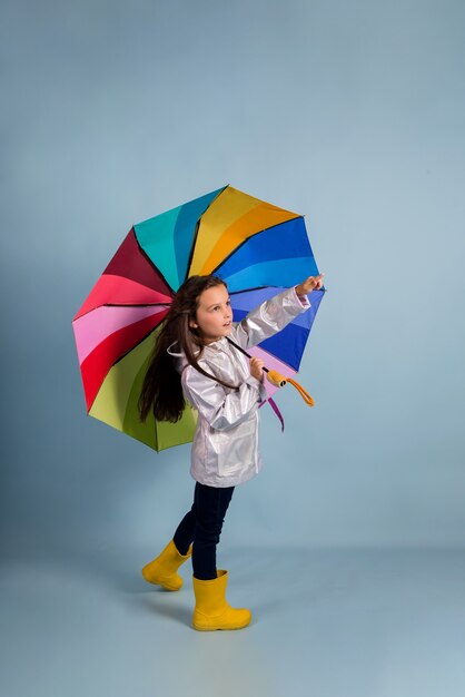 Een klein brunette meisje in een regenjas en rubberen laarzen staat onder een veelkleurige paraplu op een blauwe achtergrond met een plek voor tekst