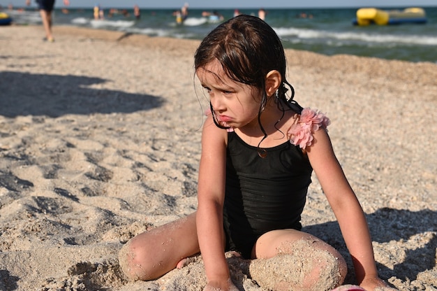 Een klein brunette meisje in een stijlvol badpak zittend op een zandstrand aan zee en spelen met het zand. Kind dat aan het zonnebaden is, geniet van een mooie zonnige dag tijdens de zomervakantie