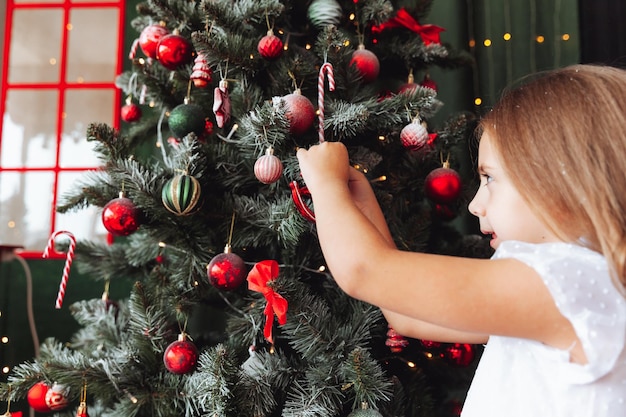 Een klein charmant meisje in een jurk hangt een glanzende bal aan de kerstboom Een kind versiert een kerstboom in een mooie woonkamer Het concept van een gelukkige familievakantie Nieuwjaar