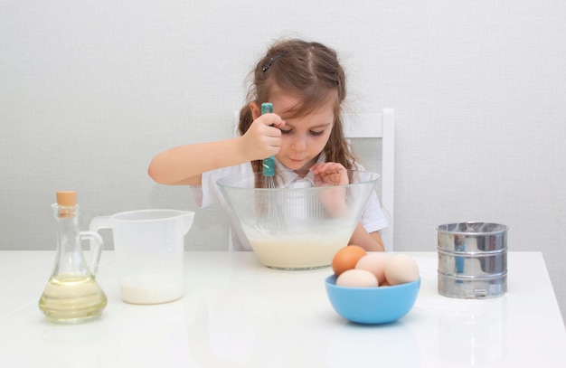 Een klein charmant meisje in een schort bereidt deeg van meel, eieren en melk, kind mengt ingrediënten in glazen kom, kleine kok bereidt voedsel. Zelfgemaakte taarten.