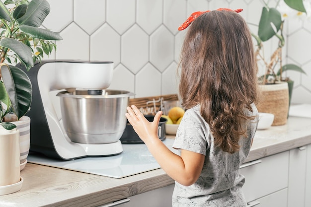 Een klein donkerharig meisje van 3 jaar bakt appeltaart in de keuken. Kind staat in de buurt van planetaire mixer. Achteraanzicht