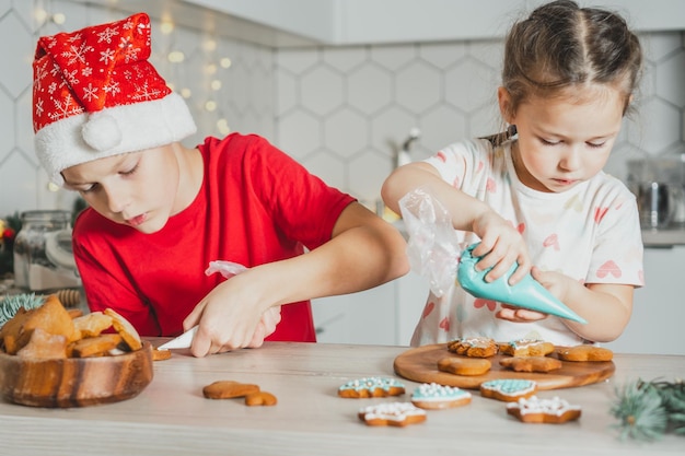 Een klein donkerharig meisje van 3 jaar en een jongen van 8 jaar in een rode kerstmuts versieren peperkoekkoekjes met glazuur. Broers en zussen in witte kerst ingerichte keuken. Vrolijk kerstfeest gelukkig nieuwjaar