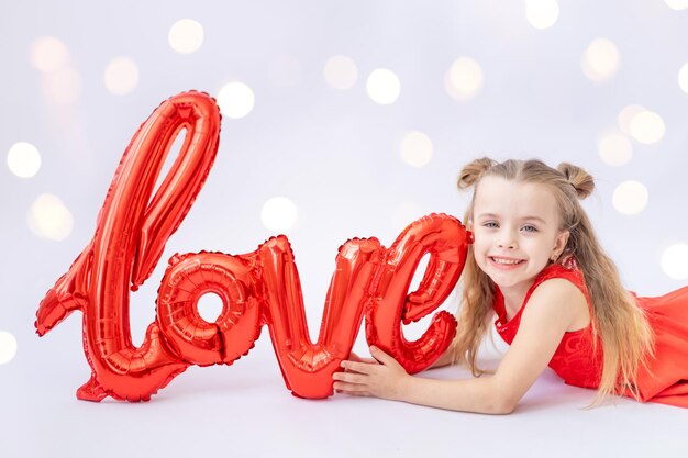 Een klein gelukkig kindmeisje in een rode jurk houdt een grote ballon vast met de inscriptie Love op een witte geïsoleerde achtergrond met een kant en lacht en glimlacht Valentijnsdag een plek voor tekst