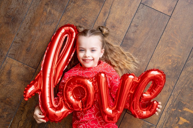 Een klein gelukkig kindmeisje in rode pyjama met een grote ballon met de inscriptie Love ligt op een donkerbruine houten achtergrond op de vloer en lacht om het concept van Valentijnsdag, een plek voor tekst