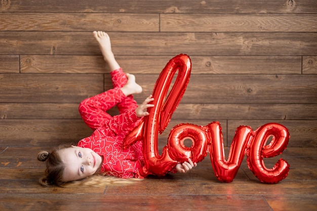 Een klein gelukkig kindmeisje in rode pyjama met een grote ballon met de inscriptie Love ligt op een donkerbruine houten achtergrond op de vloer en lacht om het concept van Valentijnsdag een plek voor tekst