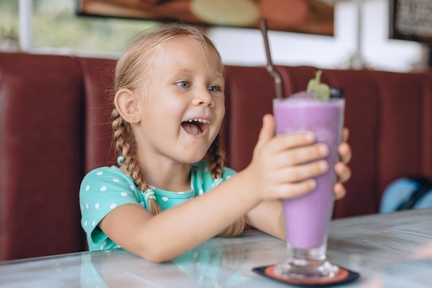 Een klein grappig kind met blonde staartjes kijkt naar een groot glas milkshake en geniet van een zoet dessert in een lokaal gezellig café. Portret.