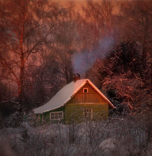 Een klein houten huis in het bos in de winter bij zonsondergang, de kachel staat aan en de rook komt uit de buis