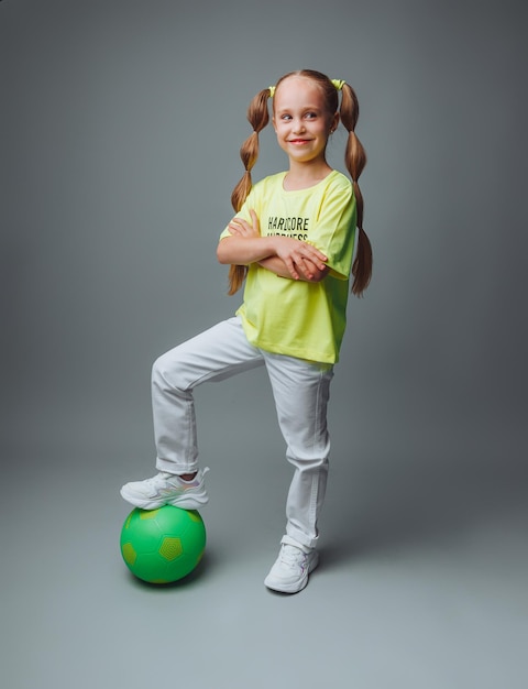 Een klein jong meisje met een groen zwaard in haar hand glimlacht naar de camera geïsoleerd op een grijze achtergrond een kleine atleet met een bal kindersport