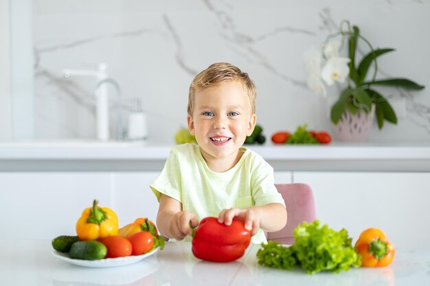 Een klein jongenskind zit aan een tafel met groenten in de keuken thuis met peper in zijn handen de baby glimlacht het concept van gezond eten vegetarisch