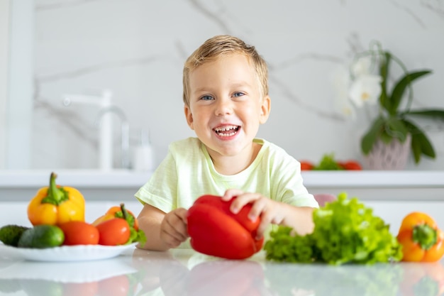 Een klein jongenskind zit aan een tafel met groenten in de keuken thuis met peper in zijn handen de baby glimlacht het concept van gezond eten vegetarisch