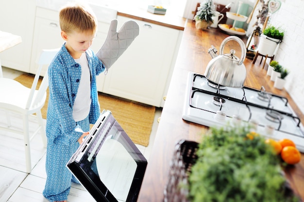 Een klein kind een jongen in blauwe pyjama met een handschoen staat tegen de achtergrond van een open oven en lacht tegen de achtergrond van de keuken