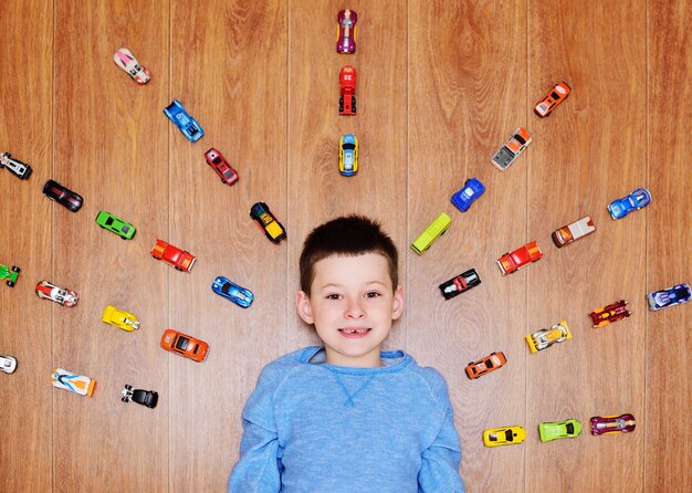 Een klein kind een jongen in een blauwe trui lacht liggend op de houten vloer tegen de achtergrond van veel speelgoedauto's.