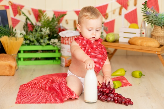 Een klein kind, een jongen van zes maanden oud, zit op de keukenvloer met een fles melk en druiven