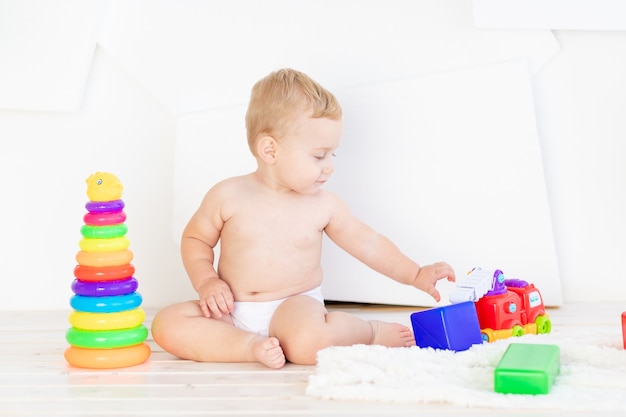 Een klein kind, een jongen van zes maanden, speelt met heldere blokjes en een piramide in een helderwitte kamer in luiers