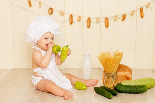 Een klein kind, een meisje van zes maanden oud, zit in de keuken met een koksmuts en een schort en eet groene paprika's