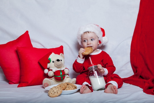 Een klein kind in een rode kerstmuts eet koekjes en melk. Kerst portret van een baby in een rode kerstmuts.