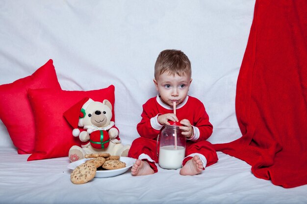 Een klein kind met een rode muts eet koekjes en melk Kerstfotografie van een baby met een rode muts Nieuwjaarsvakantie en Kerstmis