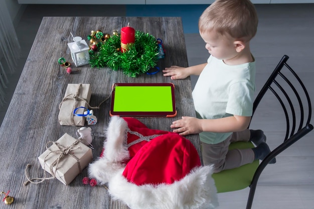 Een klein kind met kerstcadeautjes zit aan een houten nieuwjaarstafel met een tablet in zijn handen