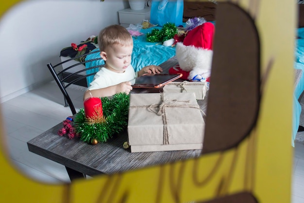 Een klein kind met kerstcadeautjes zit aan een houten nieuwjaarstafel met een tablet in zijn handen