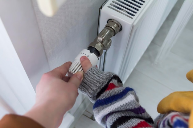 Een klein kind met zijn moeder warm gekleed in een trui en een hoed draait de kraan op de radiator Het concept van crisis en gebrek aan verwarming in huizen