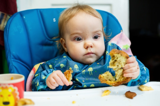 Een klein kind zit in een voedingsstoel en eet voor het eerst brood. Gezicht in kruimels