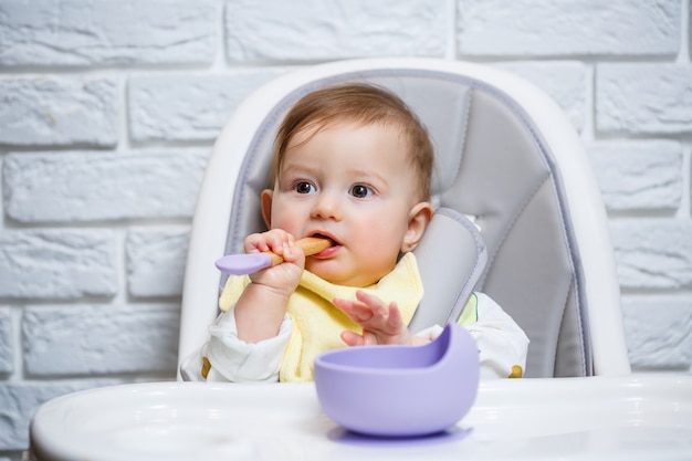 Een klein kind zit op een kinderstoel en eet voedsel van een bord met een lepel. siliconen keukengerei voor baby's om baby's te voeden