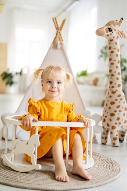 Een klein kindmeisje in een gele jurk speelt thuis met een giraf met een wigwam in een lichte kamer en lacht gelukkige jeugd