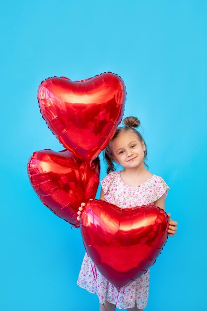 Een klein kindmeisje op een blauwe geïsoleerde studioachtergrond met rode ballonnen in de vorm van een hart heeft plezier en houdt voor de gek Ruimte voor het kopiëren van tekst Valentijnsdagconcept