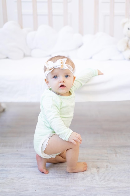 Een klein kindmeisje staat of klimt op de wieg in de kinderkamer in een wit katoenen rompertje