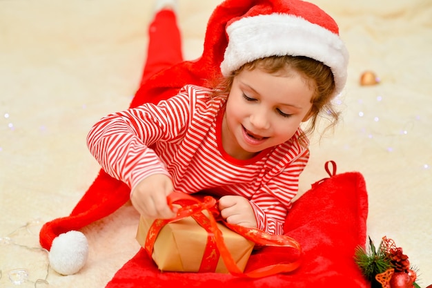 Een klein lachend meisje in de rode hoed van de kerstman en rode kerstkleding met een cadeau Nieuwjaar Kerstmis