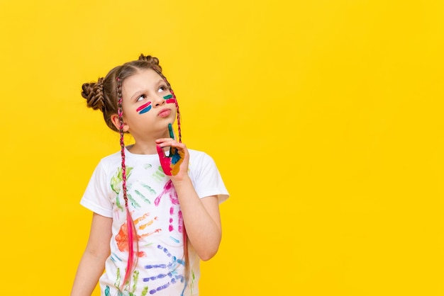 Een klein meisje beschilderd met verf kijkt naar uw advertentie op een gele geïsoleerde achtergrond De kunst van het schilderen met verf voor kinderen Kunstcursussen voor schoolkinderen Ruimte kopiëren