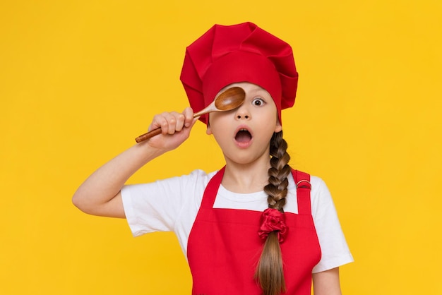 Een klein meisje chef speelt met houten lepels Koken voor kinderen Een kind in een rood schort en een pet op een gele geïsoleerde achtergrond