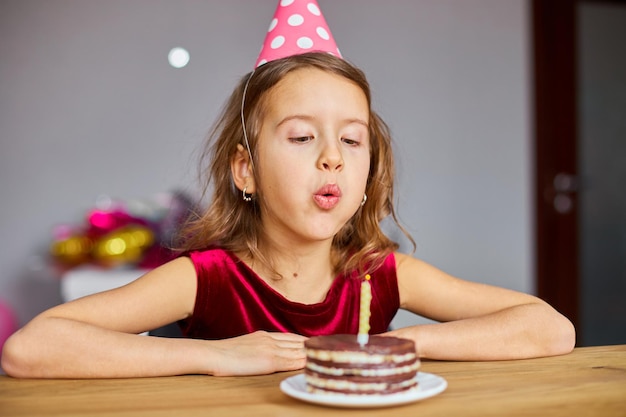 Een klein meisje draagt een verjaardagshoed terwijl ze naar een verjaardagstaart kijkt en kaarsen blaast voor een feestconcept